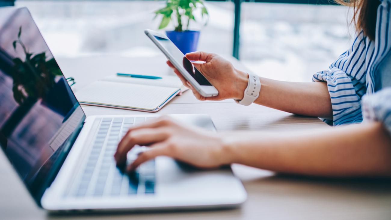 Personne écrivant sur un clavier d'ordinateur portable de la main gauche tout en ayant son téléphone en main droite